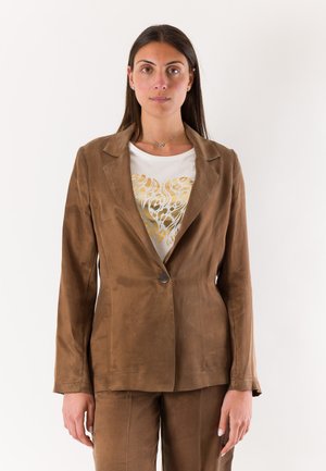 Giovane donna in piedi di fronte, indossa un blazer e pantaloni in suede marrone con una camicia bianca con un motivo a cuore dorato.