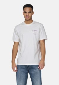 White cotton t-shirt with short sleeves, featuring purple text reading "SUNDAYS" and graphic details on the left chest area.