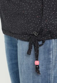 Mörkgrå topp med små prickar, utrustad med dragsko i nederkant och tre färgade pärlor. Matchad med ljusblå denimjeans.