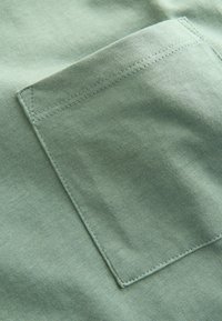 Grüner Baumwollstoff mit einer großen, rechteckigen Tasche und sauberem Stich. Die Textur wirkt weich und glatt ohne Muster.