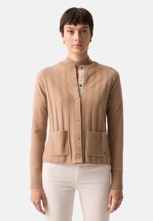 Frau trägt eine beige gerippte Strickjacke mit zwei Fronttaschen über einem hellen Top und weißen Hosen, sie steht vor einem schlichten Hintergrund.