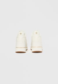 Zapatillas blancas de cuero con suela gruesa, con un detalle en oro en el talón y un logo circular en la parte trasera.
