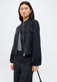 Mlada ženska, oblečena v črno bomber jakno preko črno-bele črtaste majice, temne denim hlače, stoji ob navadni ozadju.