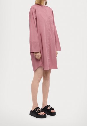 Person iført en løstsiddende, langærmet pink skjortekjole og sorte platformssandaler med dobbelte spænder, stående mod en ensfarvet baggrund.