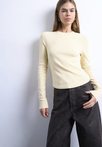 Γυναίκα με ίσια ανοιχτό καφέ μαλλιά που φοράει κρεμ μακρυμάνικη ριπ μπλούζα και σκούρα καφέ υφασμάτινη παντελόνα φαρδιάς γραμμής, ποζάροντας με το ένα χέρι στην τσέπη.