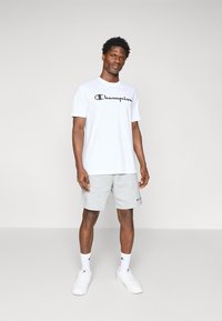 Weißes T-Shirt mit kurzen Ärmeln und einem schwarzen "Champion"-Logo, kombiniert mit grauen Shorts. Das Outfit hat eine lässige Passform und einfarbige Farben.