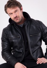 Black leather jacket with quilted shoulder accents, two zippered chest pockets, and a front zipper. Worn over a black hoodie.