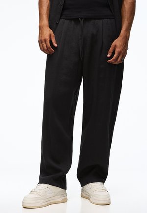 Man draagt losse zwarte broek gecombineerd met witte sneakers, staand tegen een effen witte achtergrond.