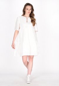 Witte jurk tot de knie met korte mouwen, details van een strik aan de voorkant, een gelaagde rok en een subtiel textuurpatroon; gedragen met witte sneakers.