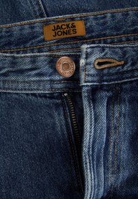 Traperice srednje plave boje s bakrenim gumbom i patentnim zatvaračem. Karakteristike uključuju narančasti šav i smeđu oznaku s natpisom "JACK & JONES."