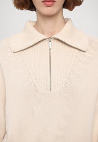 Gros plan d'une personne portant un pull en maille couleur crème avec un grand col et une fermeture éclair argentée partiellement fermée au niveau de l'encolure.