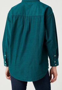 Camicia di velluto a coste color turchese con una piega sul retro, colletto a punta e polsini con bottoni. Texture liscia con orlo arrotondato e sottili righe verticali.