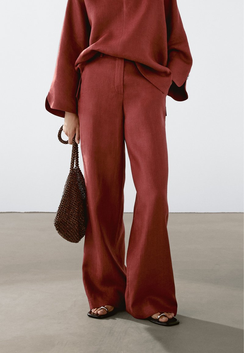 Osoba w luźnych, rdzawej kolorze szerokich spodniach i dopasowanej długiej bluzce, trzyma brązową torebkę z koralikami, stojąca na szarym podłożu.