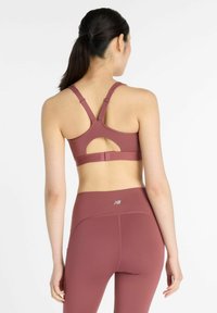 Nainen, jolla on tumma poninhäntä, pukeutuneena mauven väriseen urheiluliiviin, jossa on avoin selkäosa, ja yhteensopiviin korkeavyötäröisiin legginsseihin, seisoo valkoista taustaa vasten.