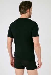 Camiseta de algodón negra de manga corta con cuello redondo. El modelo muestra la parte de atrás, vistiendo unos pantalones cortos negros ajustados. Textura suave, diseño sencillo.