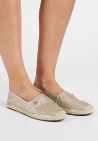 Zapatillas beige de lona perforada sin cordones con suelas de yute tejido y punta redondeada. Un detalle del logotipo circular es visible en el lateral.