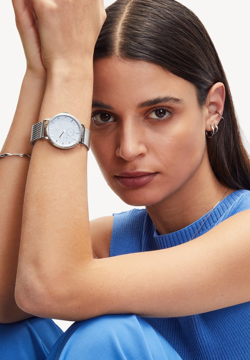 Silberne Armbanduhr mit rundem Ziffernblatt und Mesh-Armband. Das Ziffernblatt zeigt minimalistische Stundenmarkierungen und eine Datumsfunktion. Hauttöne und -texturen sind sichtbar.