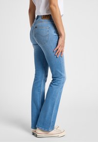 Jean évasé bleu clair à taille haute, doté d'une étiquette en cuir marron et de poches arrière. Texture de denim douce avec une coupe classique.