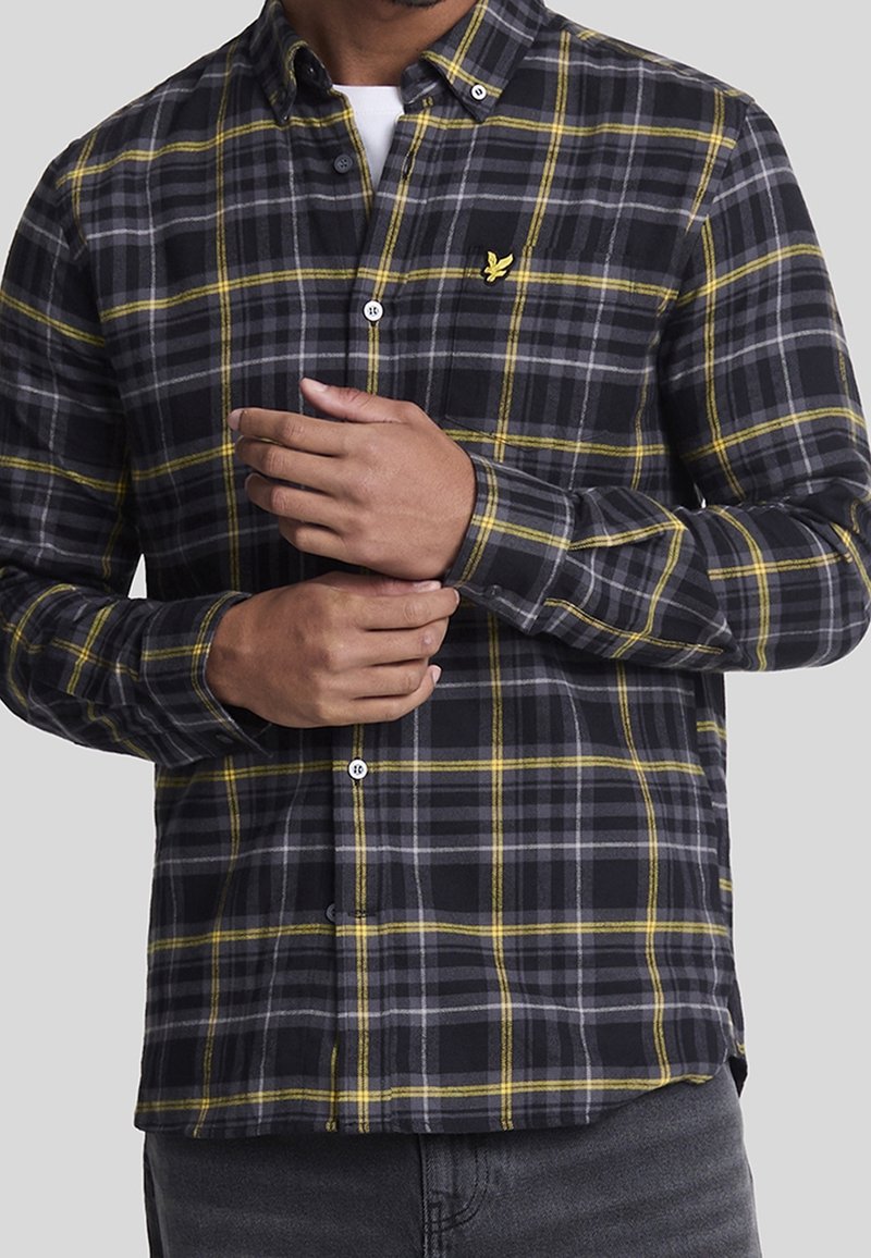Homme ajustant la manche d'une chemise à carreaux noire, jaune et grise avec un logo d'oiseau jaune sur la poitrine, portée sur un sous-vêtement blanc.