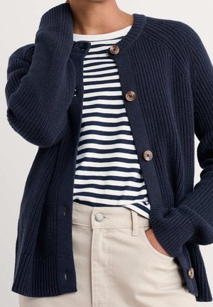 Osoba ubrana w granatowy, grubokarbowany sweter-kardigan na guziki, na biało-granatowej koszuli w paski, w połączeniu z beżowymi spodniami, z ręką w kieszeni.