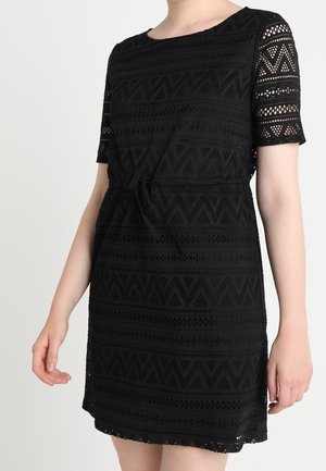 Femme portant une robe en dentelle noire avec manches courtes, col rond et ceinture nouée à la taille, visible du cou au-dessus des genoux.
