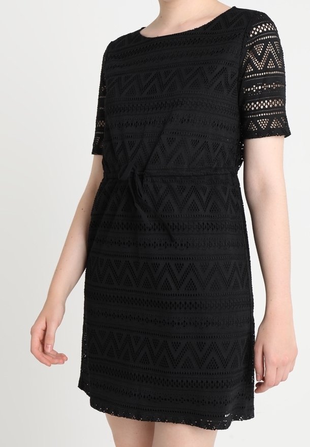 Femme portant une robe en dentelle noire avec manches courtes, col rond et ceinture nouée à la taille, visible du cou au-dessus des genoux.