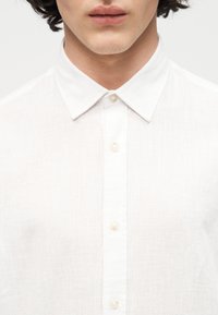 Κοντινή λήψη ενός ατόμου που φοράει λευκό πουκάμισο με κουμπιά και μυτερό γιακά, εμφανίζοντας τα χείλη, το πηγούνι και την περιοχή του πάνω μέρους του στήθους.