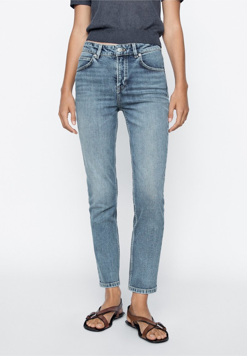 Jean en denim bleu clair avec une coupe droite, design à cinq poches et léger effet froissé. Associé à des sandales noires.