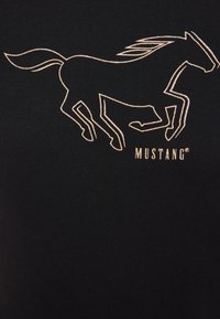 Čierne tričko s vyšívaným siluetou bežiaceho koňa v zlatom orámovaní a slovom "MUSTANG" v zodpovedajúcom zlatom písmene nižšie.