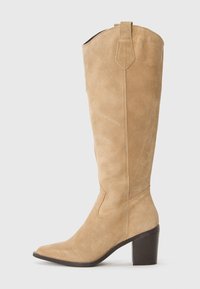Botte en suède beige jusqu'au genou avec un bout pointu et un talon épais. Présente des accents de couture et une languette sur le dessus.