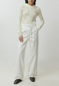 Witte gebreide top met lange mouwen en opengewerkte patroon, gecombineerd met hoge witte broeken en zwarte platte schoenen.