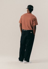 Άνδρας που φοράει ένα κόκκινο και μπεζ ριγέ πουκάμισο, μαύρο παντελόνι, λευκά αθλητικά παπούτσια και μαύρο καπέλο, στέκει με τα χέρια στις τσέπες, αντικρίζοντας μακριά.