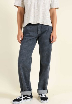 Personne portant un jean bleu foncé à motifs, retroussé aux chevilles, des baskets blanches et noires, et une chemise à manches courtes beige clair avec un motif subtil.