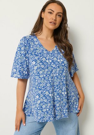 Kvinde med langt brunt hår iført en blå blomsterprintet bluse med korte ærmer og V-hals samt lyseblå jeans, stående foran en ensfarvet baggrund.