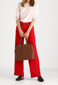 Brown leather handbag with a rounded shape, gold-tone hardware, and double handles; model wears a light top and red wide-leg pants.