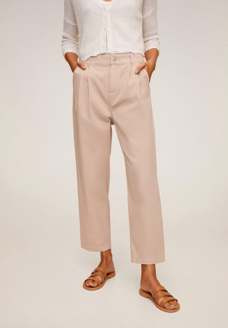 Kobieta w beżowych, szerokich, krótkich spodniach, białym szydełkowym kardiganie i brązowych klapkach, stoi z rękami w kieszeniach.