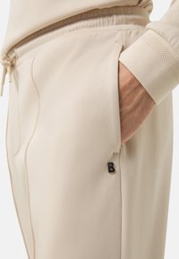 Beige Jogginghosen mit einem Kordelzugbund, seitlichen Taschen und einem kleinen schwarzen Logo-Detail. Der Stoff hat eine weiche, glatte Textur.