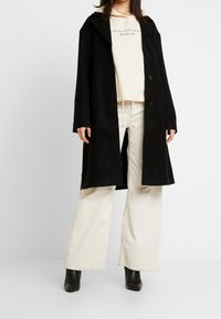 Svart ullkapp med spetsad krage, tre knappar och en avslappnad passform. I kombination med krämfärgade kordfläskbyxor och svarta ankelboots.