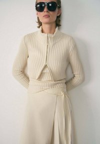 Beige ribbed sweater with a high collar and front zipper, paired with a light beige wrap skirt featuring pleats and a side tie detail.