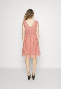 Vestido rosa con un cuerpo de encaje, diseño de espalda en forma de V y una falda de tul fluida y en capas. Lleva sandalias negras de tacón bloque.