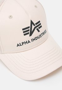 Beige baseball cap med buet skygge, med en svart logo og teksten "ALPHA INDUSTRIES" på forsiden, netting-sider og broderte detaljer.