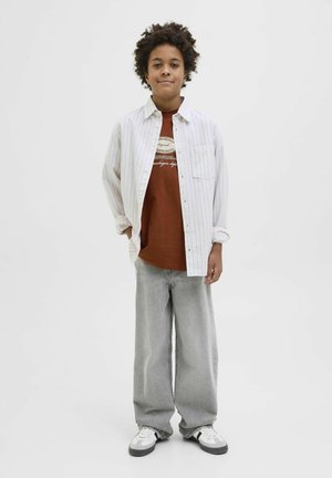 Witte gestreepte overhemd met knopen boven een bruine graphic t-shirt, gecombineerd met losse grijze jeans en zwart-witte sneakers. Witte achtergrond.