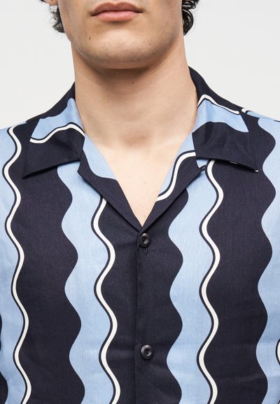 Άνδρας με πουκάμισο με κουμπιά, με κυματιστή κάθετη ρίγα σε σκούρο και ανοιχτό μπλε χρώμα και ανοιχτό γιακά.