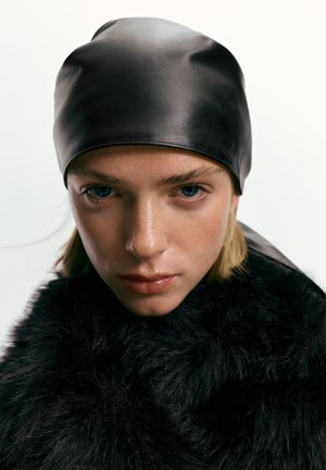Close-up of person with blue eyes wearing a black leather cap and a thick black fur coat against a white background.