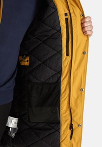 Giacca esterna gialla con interno trapuntato nero, dotata di tasca con zip, tasca in rete e profili blu a contrasto lungo il bordo.
