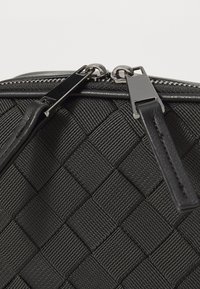 Close-up van zwarte geweven textuurstof met twee zilveren ritssluitingen en zwarte leren ritslips op een tas.