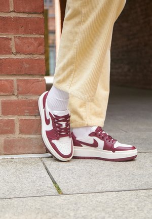 Person trägt beige Hose und weiße Socken mit rot-weißen Nike-Sneakers und steht an einer roten Ziegelwand auf einem gefliesten Boden.