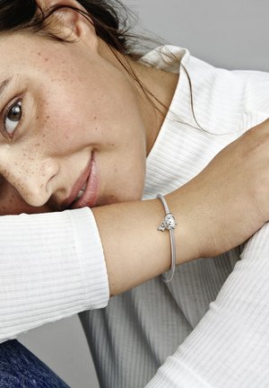 Silberne Charmarmband am Handgelenk, mit einem detaillierten Charm und einem glatten, geflochtenen Design. Die Person trägt ein weißes geripptes Oberteil.