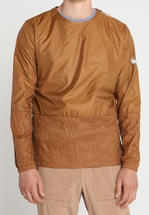 Mann trägt eine langärmelige, hellbraune Windjacke mit gesteppter Unterseite und beigen Hosen vor einem schlichten Hintergrund.