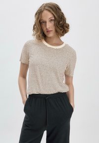 Kerman t-paita, jossa on vaalean kermanväriset ja ruskeat vaakaraitat, pyöreä kaula-aukko, yhdistetty mustien naruvarustesateenkaarihousujen kanssa.